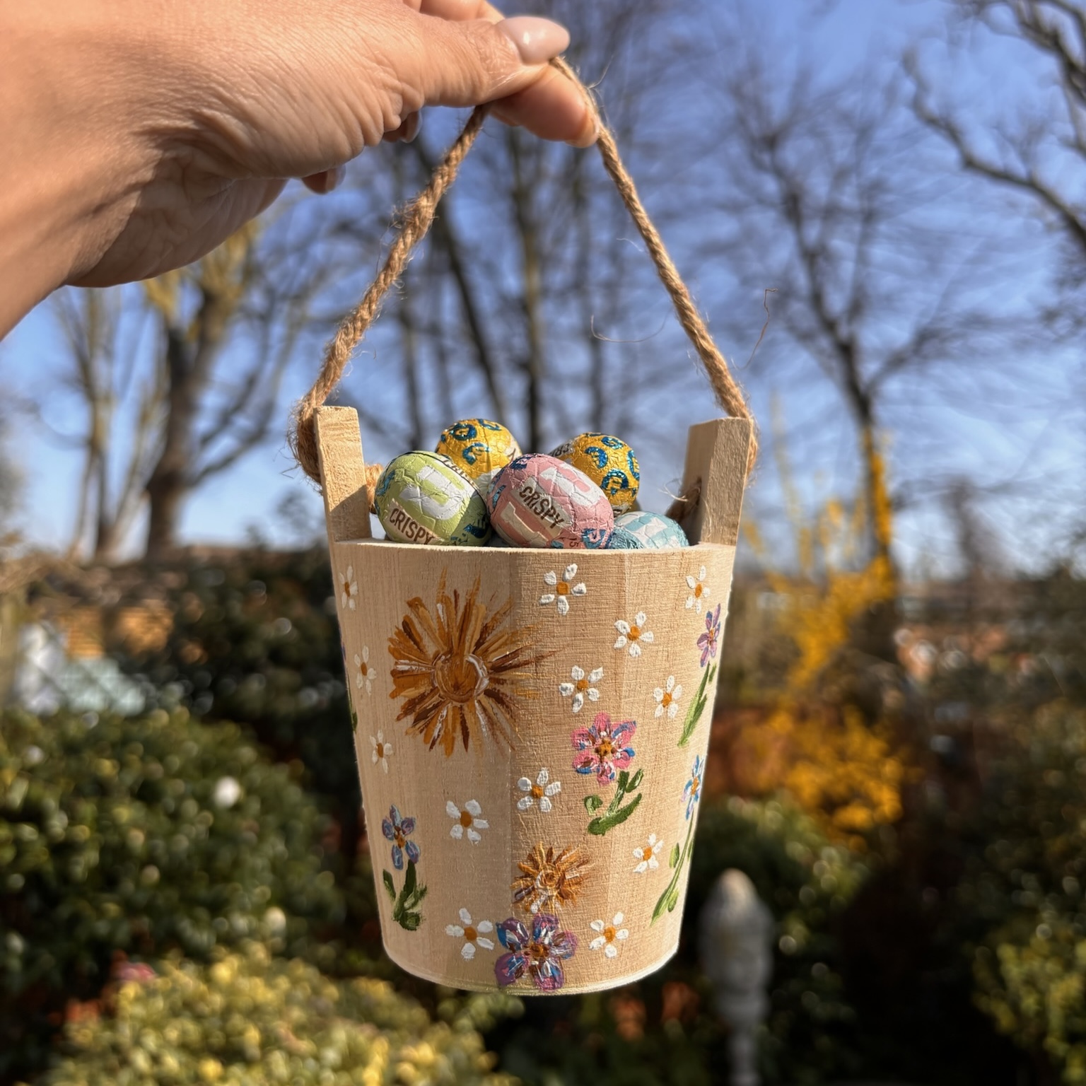 Painted wooden bucket is held up on a sunny day filled with small Easter eggs.